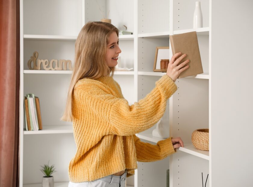 Young woman taking book from shelf at home