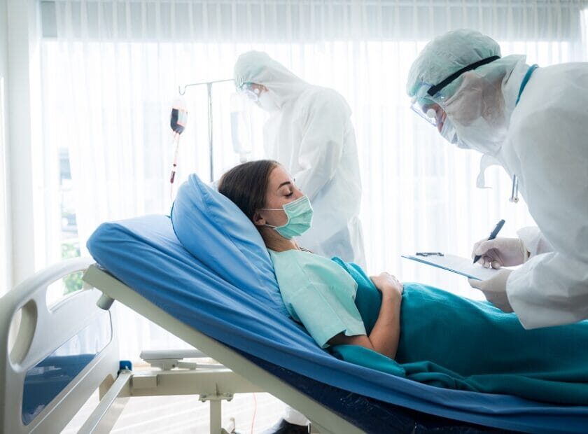 Young woman suffering with corona virus infection lying on hospital bed with mask sleeping and resting while doctor taking notes of progress and writing on clipboard in background wearing ppe kit