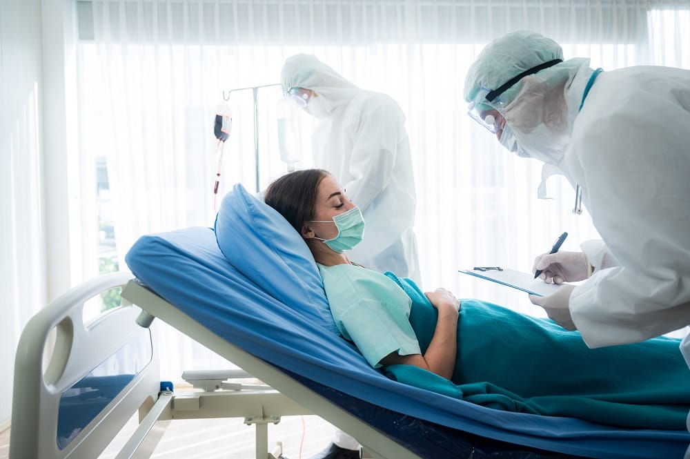 Young woman suffering with corona virus infection lying on hospital bed with mask sleeping and resting while doctor taking notes of progress and writing on clipboard in background wearing ppe kit